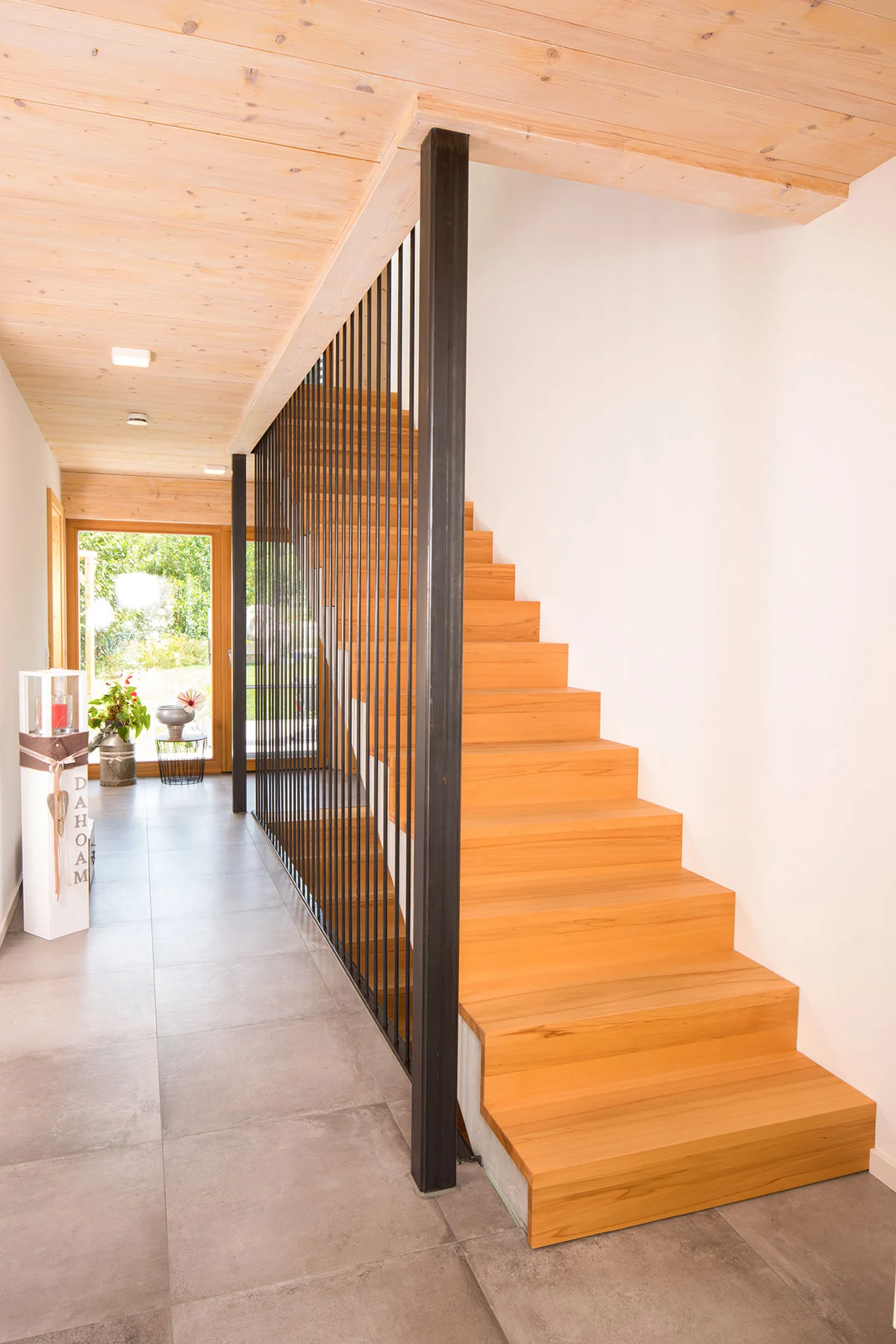 Eine moderne Faltwerktreppe aus Holz mit schwarzen Metallstäben und -balken als Geländer. Eine verputzte Wand und die Sichtholzdecke des Holzhauses sind sichtbar. © Brunthaler Naturhausmanufaktur