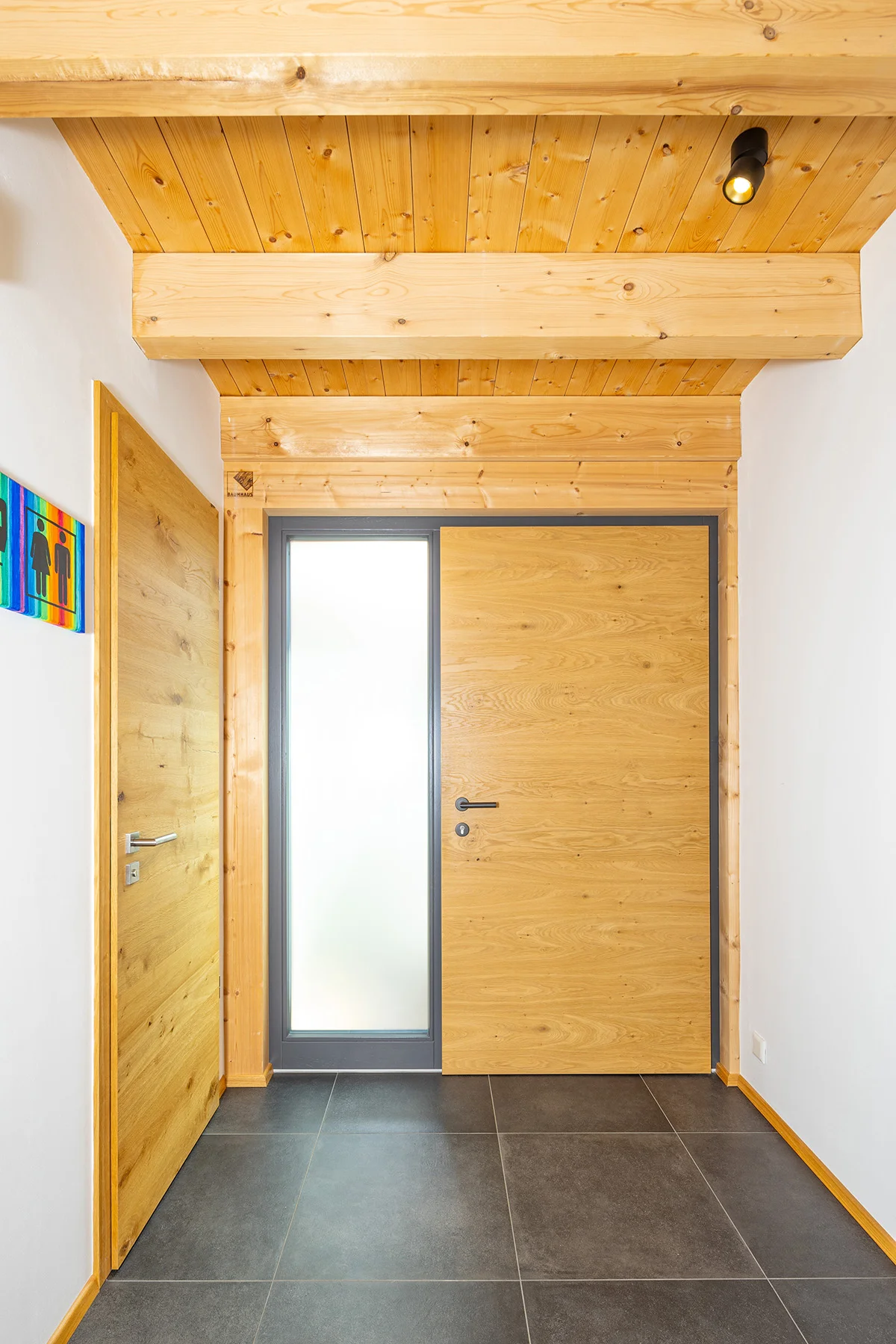 Die Eingangstür des Holzhauses in Holz mit seitlichem bodentiefen Milchglasfenster. Dunkle großen Fliesen am Boden und eine Holzdecke. © Brunthaler Naturhausmanufaktur