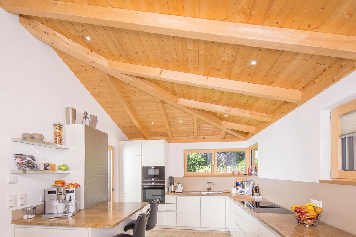 Eine geräumige helle Küche mit Frühstückstheke, Kaffeemaschine, ein paar Barhocker. Im Fokus ist die Holzdecke mit eingesetzten Licht-Spots, die Balken des Dachstuhls sind wie Adern eines Blattes Richtung hinterer Ecke des Erkerraums gefächert. © Brunthaler Naturhausmanufaktur