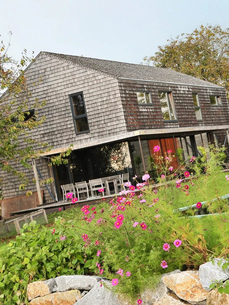 Modernes Massivholzhaus mit vergrauter Holzschindelfassade ohne Dachüberhang. Davor sieht man einen grünen Rasen mit einer kleinen Steinmauer und bunter Bepflanzung © Brunthaler Naturhausmanufaktur