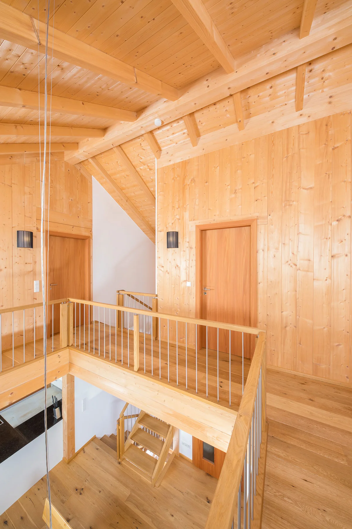 Eine Galerie im Wohnhaus, viele Wände haben eine Sichtholzoberfläche, ein paar sind verputzt. Man sieht durch die Galerie nach unten. Das Geländer ist auch aus Holz und aus Edelstahl. Man sieht ein paar Türen aus Holz und den Dachstuhl aus Sichtholz. © Brunthaler Naturhausmanufaktur