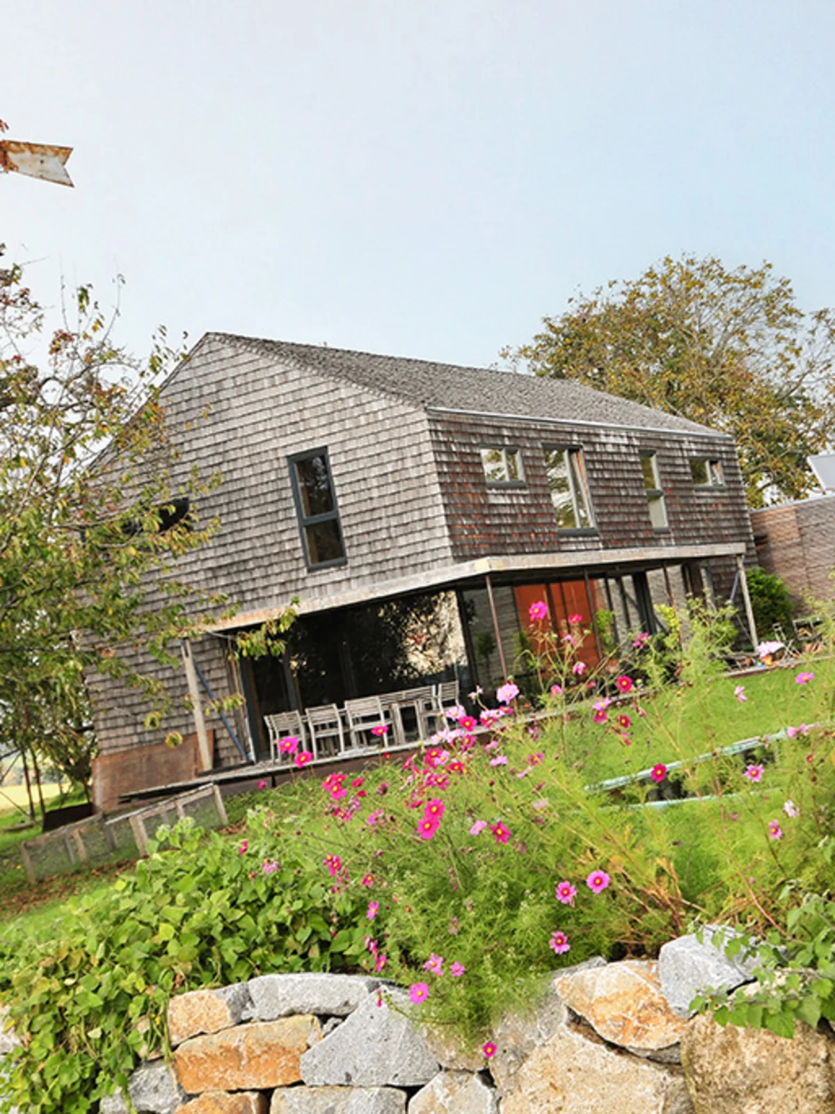 Modernes Massivholzhaus mit vergrauter Holzschindelfassade ohne Dachüberhang. Davor sieht man einen grünen Rasen mit einer kleinen Steinmauer und bunter Bepflanzung © Brunthaler Naturhausmanufaktur