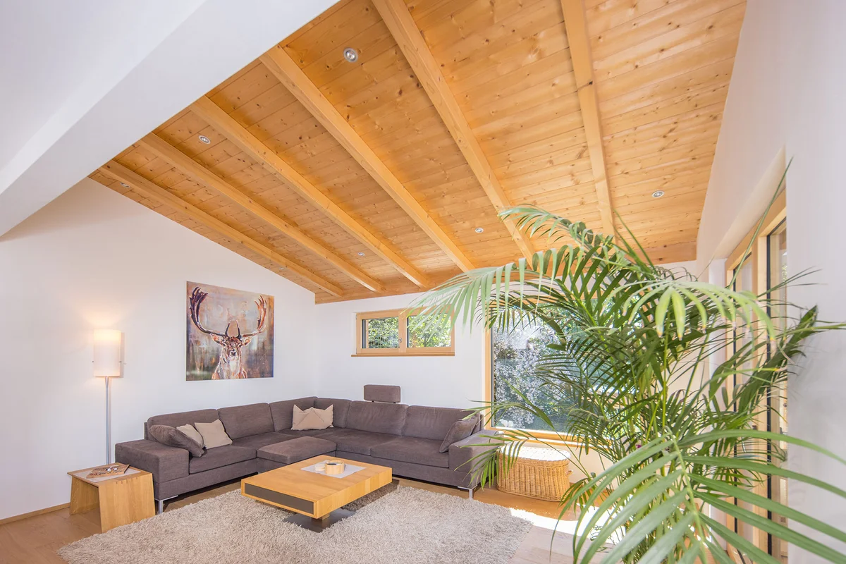 Wohnzimmer in einem modernen Holzhaus. Die warmgraue Couch und der minimalistische Couchtisch aus Holz harmonieren mit den verputzten Holzwänden und dem Sichtdachstuhl. © Brunthaler Naturhausmanufaktur