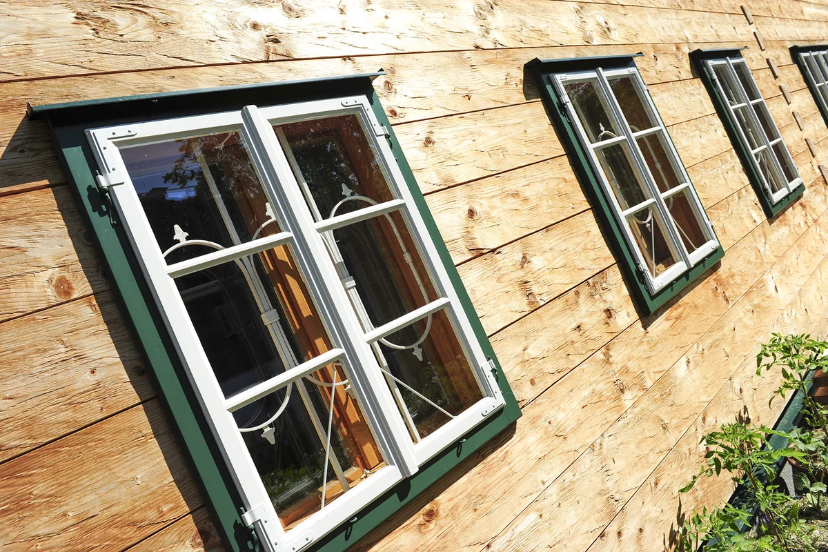 Detailaufnahme einer Blockbohlenfassade mit grün gerahmten Fenstern und weißen Fensterläden. © Brunthaler Naturhausmanufaktur