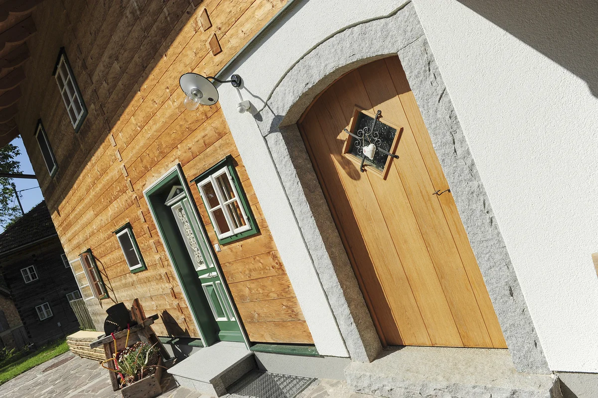 Zwei urige Türen, eine grün mit filigranen weißen Verzierungen und eine aus Holz umrandet mit Stein und einem kleinen Fenster. Blockbohlenfassade eines Massivholzhauses. © Brunthaler Naturhausmanufaktur