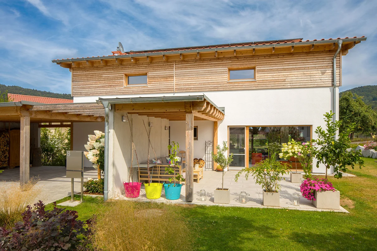 Klassisches Einfamilienhaus mit verputzter Fassade und einem kleinen Teil an horizontaler Holzfassade. Große Fenster und eine betonierte Terrasse mit Überdachung ergänzen das Holzhaus. © Brunthaler Naturhausmanufaktur