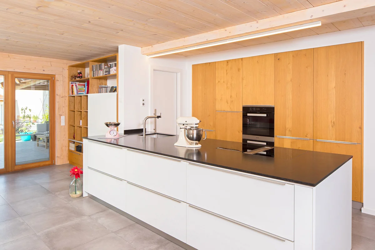 Eine moderne Küche mit ebenen Einbauschränken und einer weißen Kücheninsel mit schwarzer Arbeitsfläche. © Brunthaler Naturhausmanufaktur