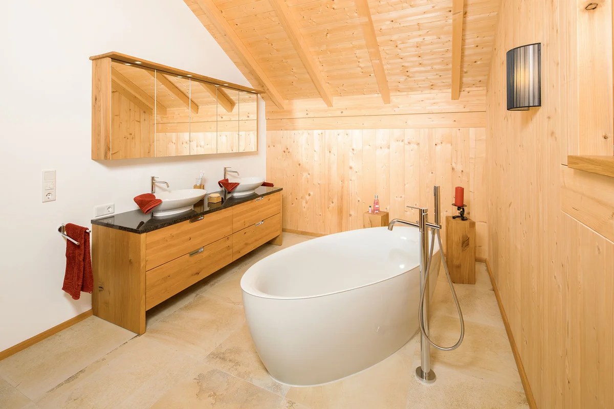 Badezimmer im Massivholzhaus mit Sichtholzwänden und verputzten Wänden. Eine freistehende Badewanne und großzügigen Waschbeckenbereichen. Man sieht den Dachstuhl aus Sichtholz. © Brunthaler Naturhausmanufaktur