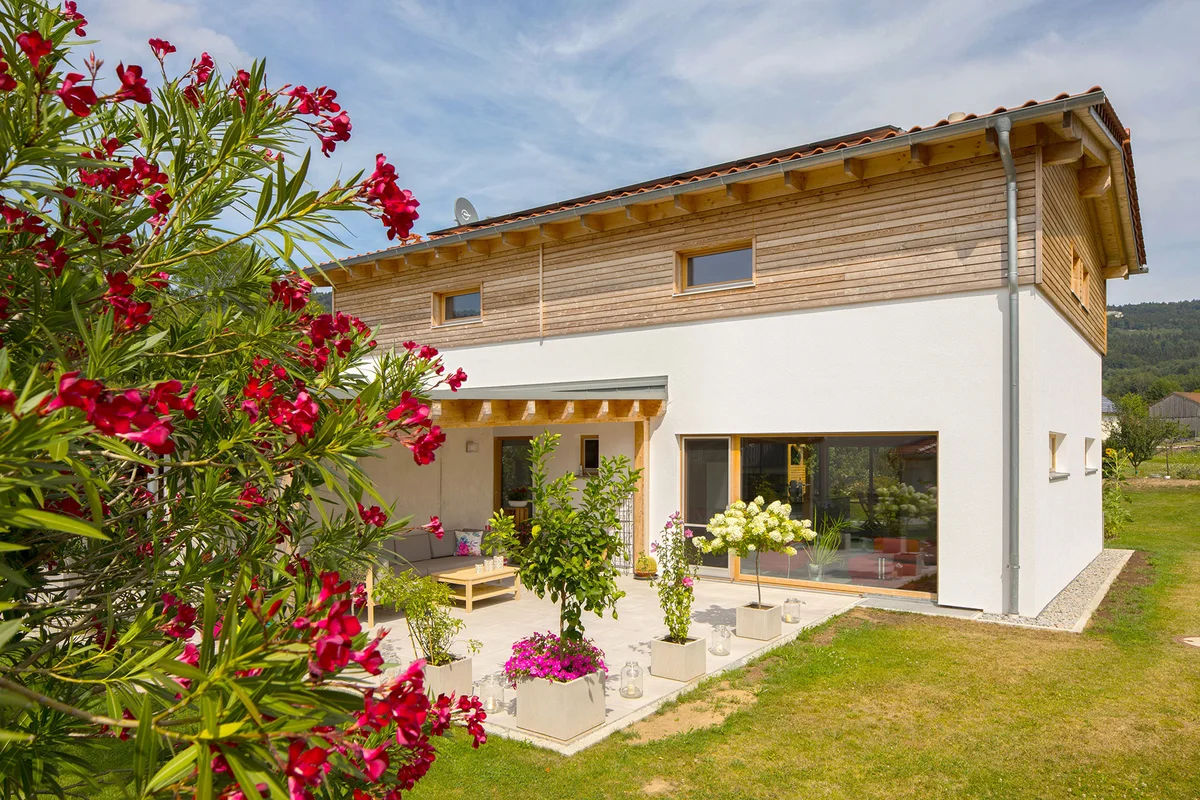 Klassisches Einfamilienhaus mit verputzter Fassade und einem kleinen Teil an horizontaler Holzfassade. Große Fenster und eine betonierte Terrasse ergänzen das Holzhaus. Im Vordergrund ist ein blühender Rhododendron. © Brunthaler Naturhausmanufaktur