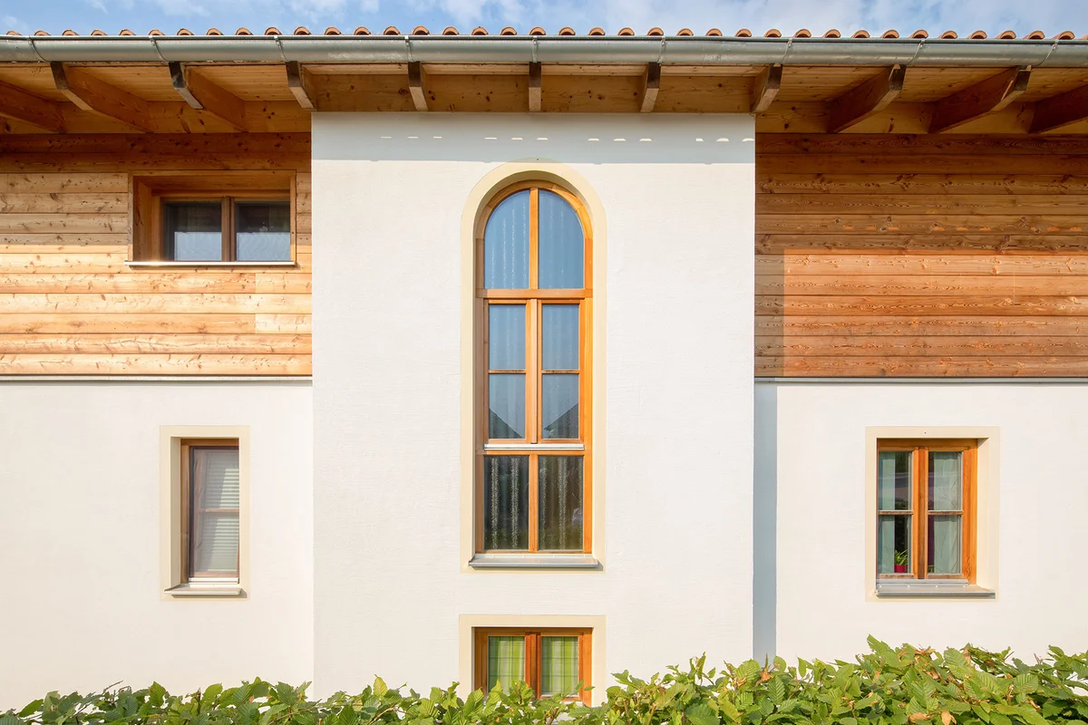 Detailaufnahme eines Erkers des halbverputzten Massivholzhaus mit abgerundetem hohem Fenster und Unterteilung durch Holzsprossen. © Brunthaler Naturhausmanufaktur