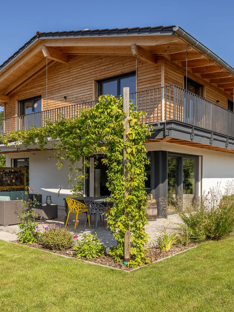 Klassische Einfamilien-Holzhaus mit umlaufendem Balkon in Hanglage. Oben Holzfassade unten verputzt. Gemütliche Terrasse mit Raumteiler aus gestapeltem Stückholz und Kletterwein, der sich zum Balkon hochrankt. © Brunthaler Naturhausmanufaktur
