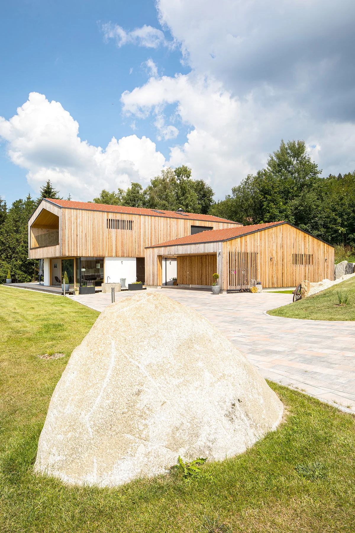 Modernes Architektenholzhaus mit vertikaler heller Holzfassade und verputztem Erdgeschoss. Ein großer überhängender Balkon und eine große Garage mit gleicher Fassade sind sichtbar. Das Haus liegt einzeln umgeben von Bäumen, Rasen und einer Einfahrt mit hellem Findling-Stein. © Brunthaler Naturhausmanufaktur
