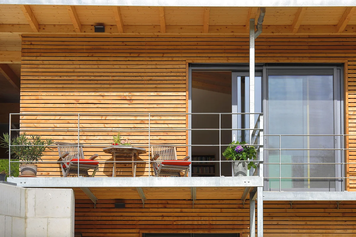 Detailaufnahme des Zugangs über den Balkon durch die Hanglage. Dekoriert mit Bestuhlung und Pflanzen. Die Balkontür ist offen. Man sieht die horizontale Holzfassade des Holzhauses. © Brunthaler Naturhausmanufaktur
