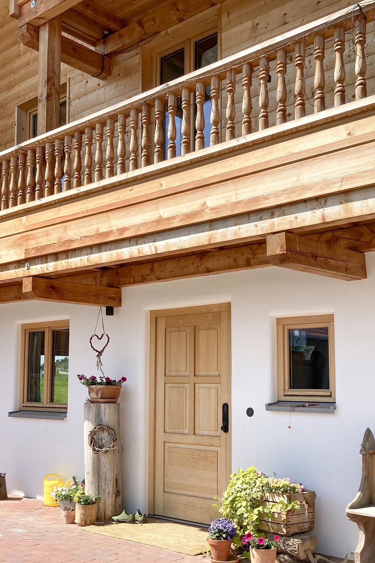 Detailansicht der Front eines bayerischen Landhauses mit urigem Holzbalkon aus gedrechselten Geländerpfosten. Die helle Holztür im unteren verputzten Geschoss harmoniert mit der Blockbohlenfassade in den oberen Stockwerken. © Brunthaler Naturhausmanufaktur