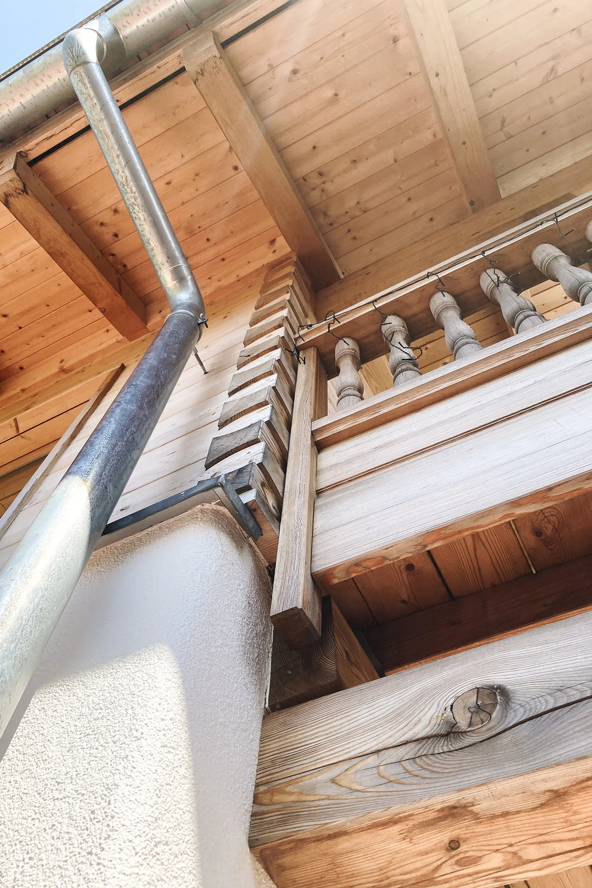 Detailaufnahme eines bayerischen Landhauses, perspektivisch von Unten. Man sieht die untere verputzte Wand und die Blockbohlenfassade mit der Eckverbindung sowie die gedrechselten Geländerpfosten des Balkons. © Brunthaler Naturhausmanufaktur