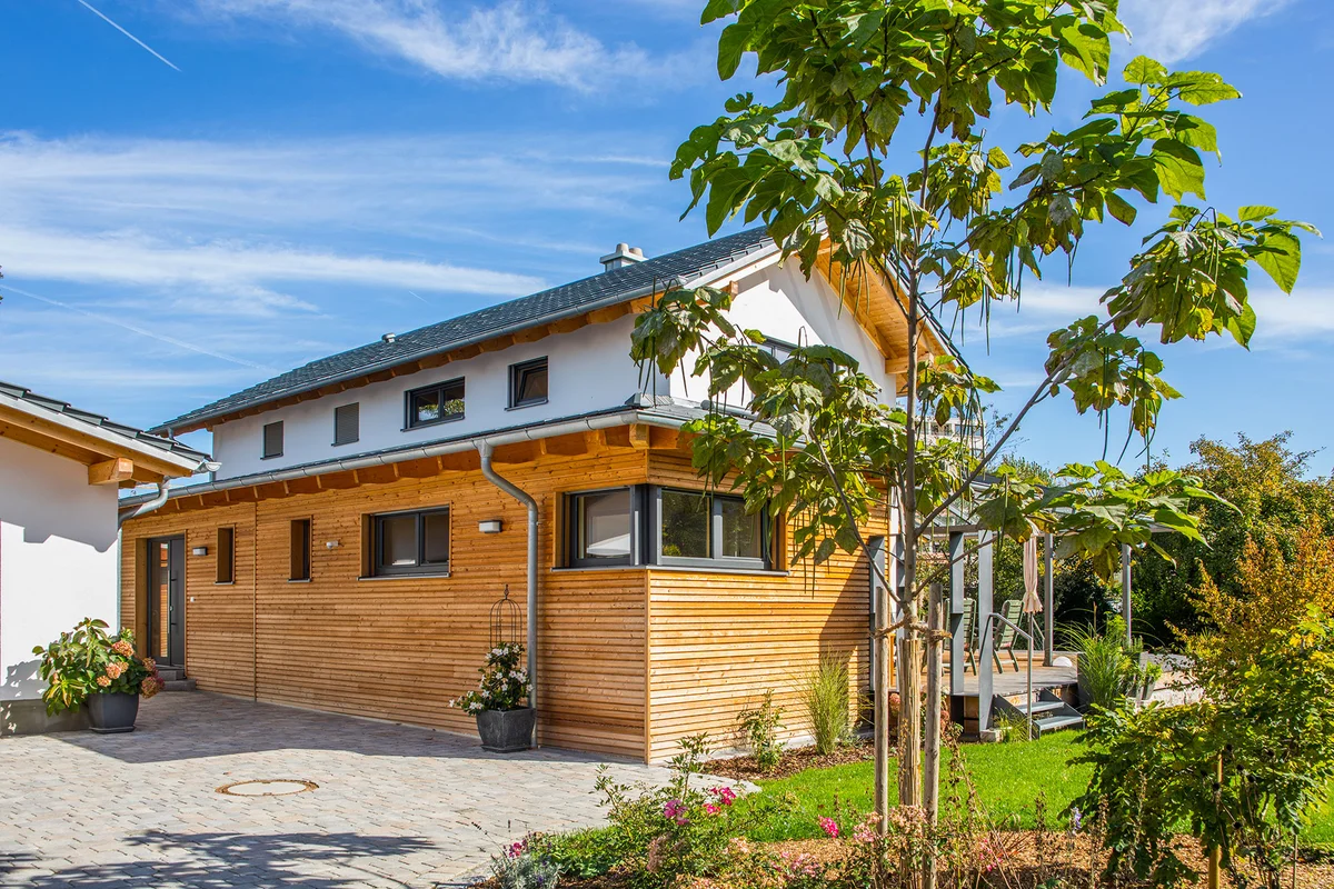Das Holzhaus ist teilweise verputzt und hat einen überdachten Übereck-Erker mit einer horizontalen Holzfassade. Ein Baum steht im Vordergrund neben der Einfahrt. © Brunthaler Naturhausmanufaktur