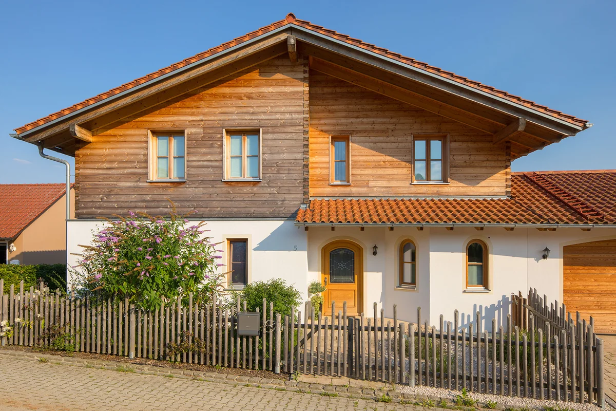 Großzügiges Massivholz-Landhaus mit Blockbohlenfassade oben und verputzter Fassade im Erdgeschoss. Rustikaler Zaun umsäumt das Grundstück. © Brunthaler Naturhausmanufaktur