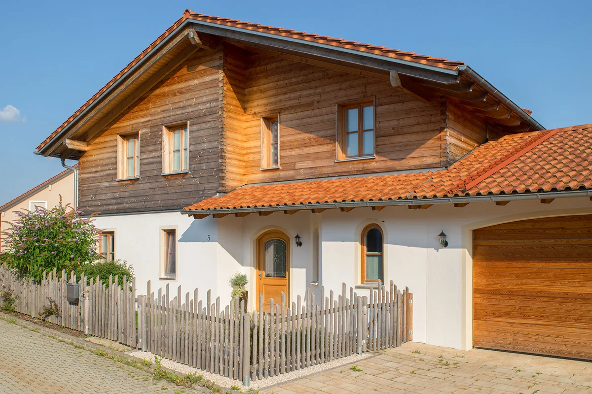 Großzügiges Massivholz-Landhaus mit Blockbohlenfassade oben und verputzter Fassade im Erdgeschoss. Rustikaler Zaun umsäumt das Grundstück. © Brunthaler Naturhausmanufaktur