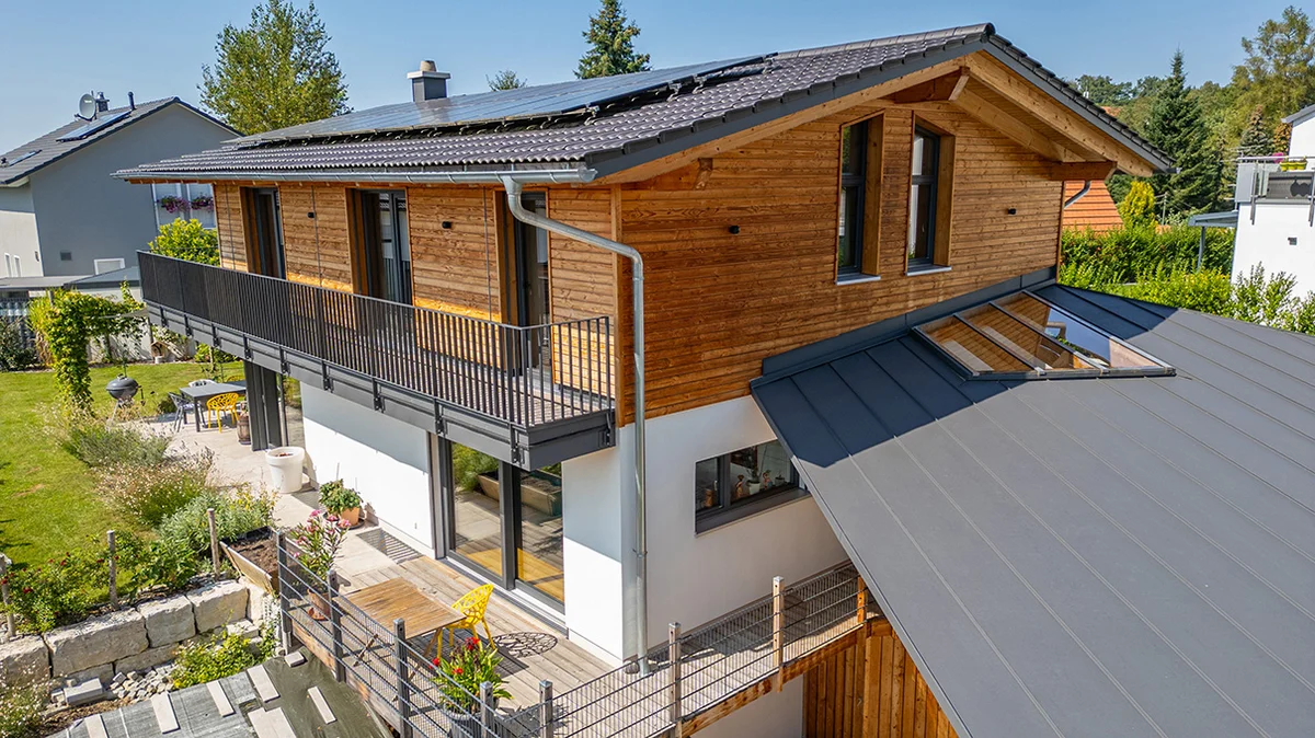 Klassisches Einfamilien-Holzhaus mit umlaufendem Balkon und dunkelgrauen Dachschindeln in Hanglage. Oben Holzfassade und unten verputzt. © Brunthaler Naturhausmanufaktur