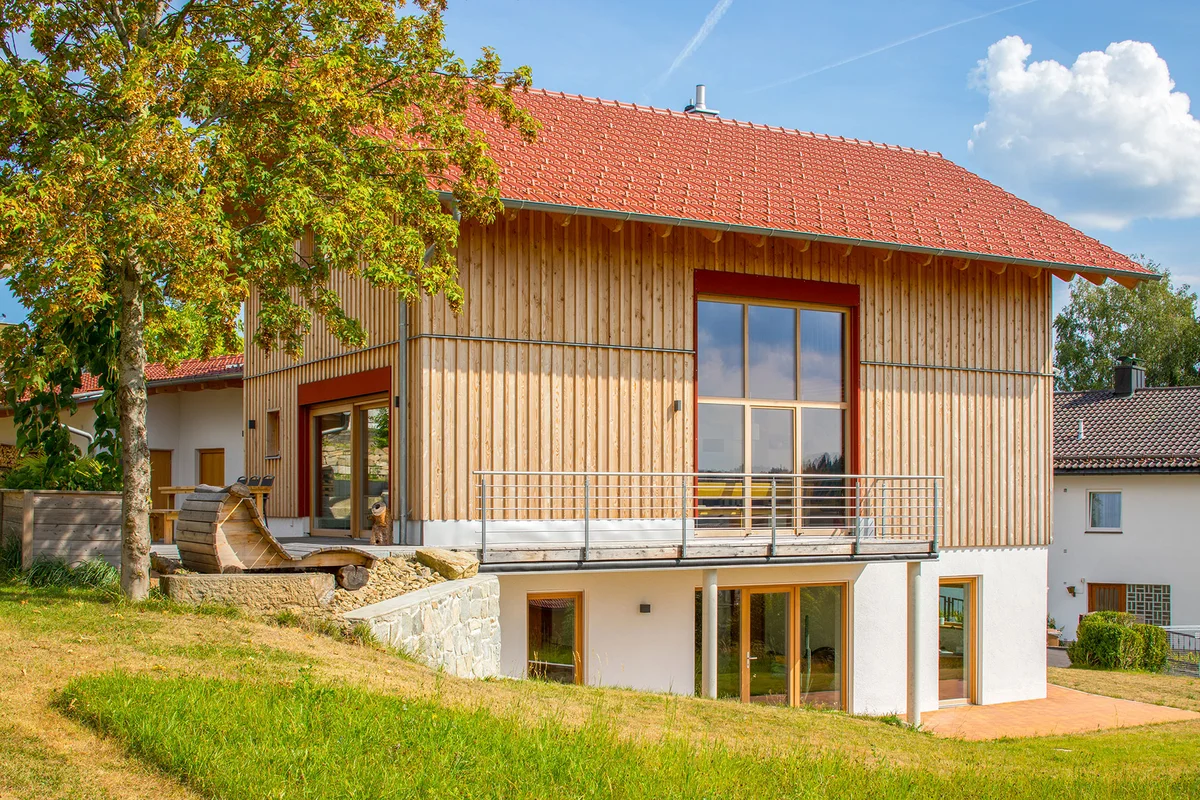 Modernes Holzhaus in Hanglage mit vertikaler Holzfassade, roten Farbakzenten, einer Holzterrasse, die sich seitlich um das Haus zieht. Der untere Bereich der Fassade ist weiß verputzt. Seitlich hat das Haus ein mehrstöckiges Fenster. Das Haus hat ein rotes Satteldach. © Brunthaler Naturhausmanufaktur