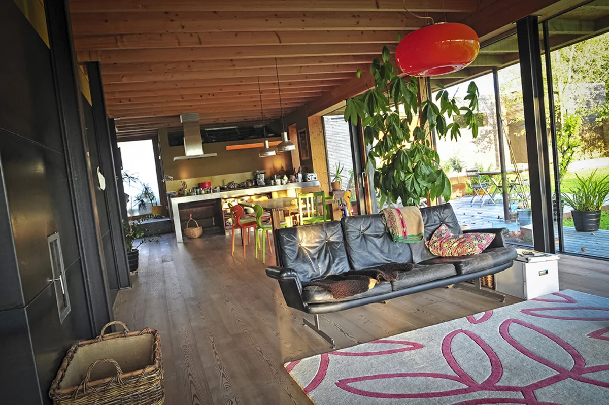 Offener Ess-, Wohn- und Küchenbereich mit großer Glasfront und Sicht auf den Garten. Links ist eine dunkle Wand mit eingebautem, zentralem Ofen. Sichtholzbalkendecke eines Massivholzhauses. © Brunthaler Naturhausmanufaktur