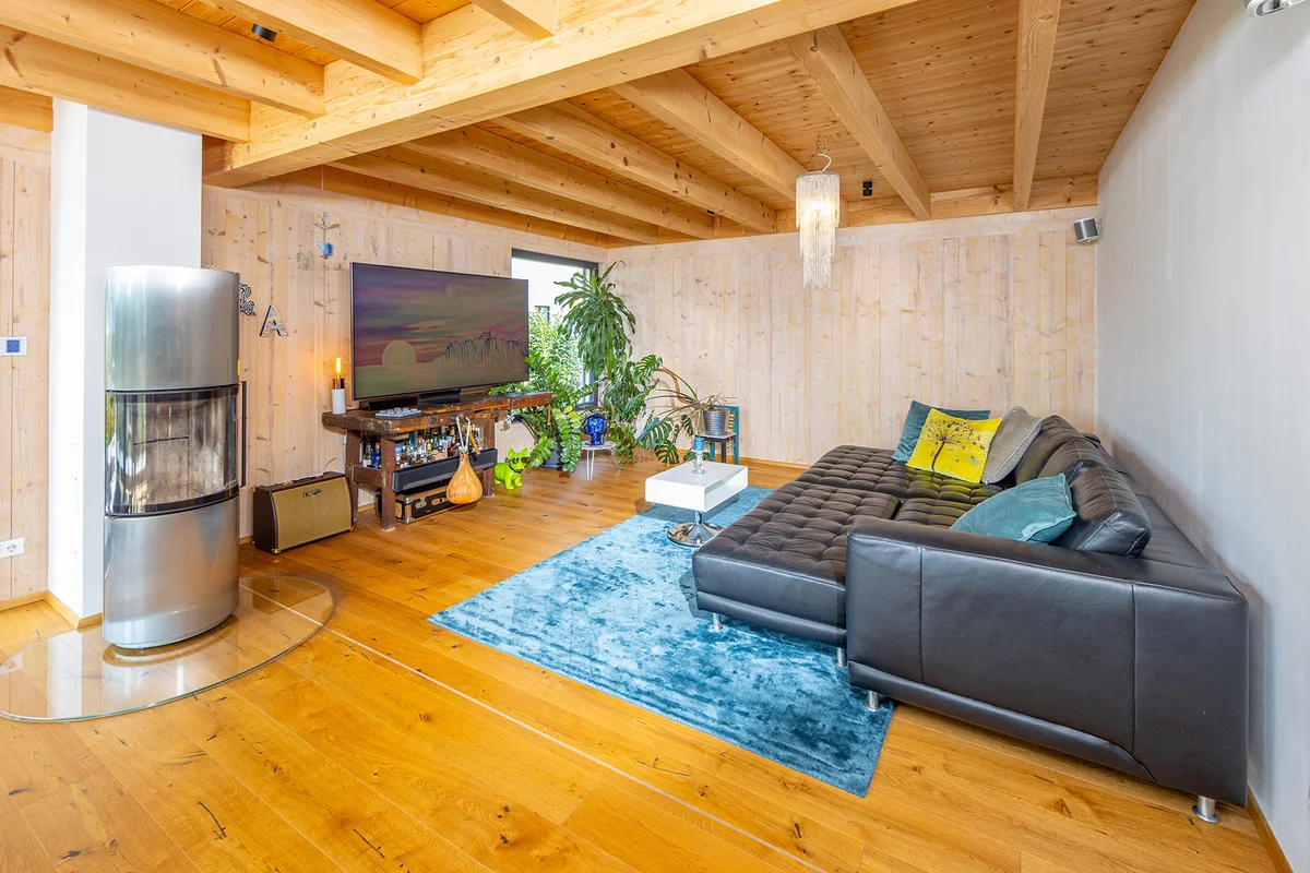 Geräumiges Wohnzimmer mit hellen Holzwänden, einem warmen Holzfußboden und einer Holzdecke mit sichtbaren Balken. Ein blauer Teppich und eine große schwarze Ledercouch stehen vor einem großen Fernseher mit einem freistehenden Holzofen aus silbernem Metall. © Brunthaler Naturhausmanufaktur