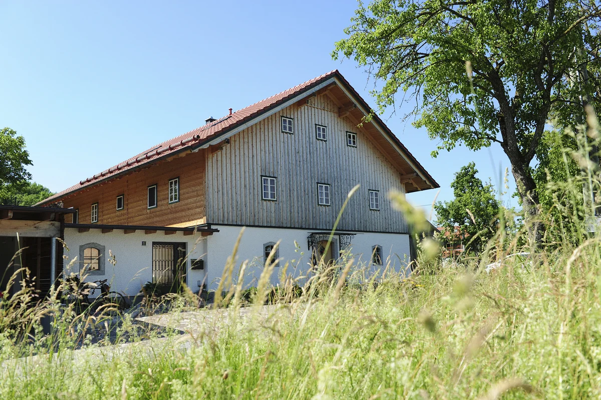 Innviertler Hof aus Massivholz mit traditioneller Holzschindelfassade, weiß verputztem Erdgeschoss und rotem Satteldach inmitten einer grünen Wiesenlandschaft. © Brunthaler Naturhausmanufaktur