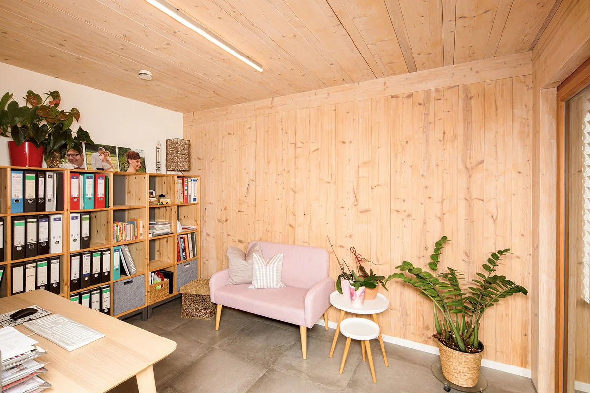 Arbeitszimmer mit einer verputzten Wand und einer Sichtholzwand, sowie Sichtholzdecke. Holzregale mit Akten und ein altrosa Sessel, sowie ein Schreibtisch sind sichtbar. © Brunthaler Naturhausmanufaktur