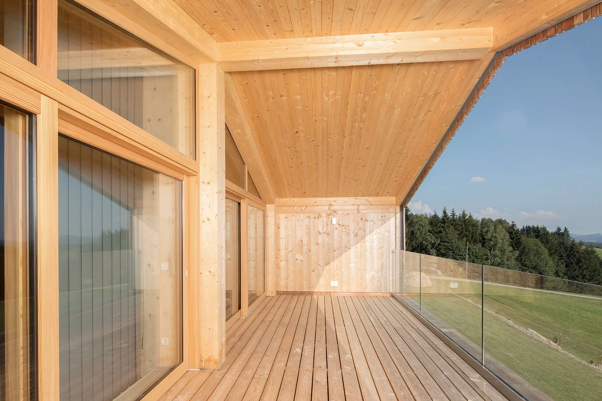 Großer, moderner, geradliniger Holzbalkon mit Glasgeländer und weiter Sicht auf die Landschaft. Auf der Hausseite ist eine große Glasfront. © Brunthaler Naturhausmanufaktur