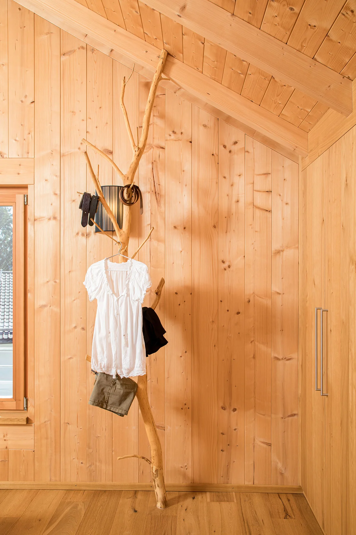 Ein Ast, der an Boden und Decke angemacht als rustikaler und kreativer Kleiderständer fungiert, steht vor einer Massivholzwand. © Brunthaler Naturhausmanufaktur