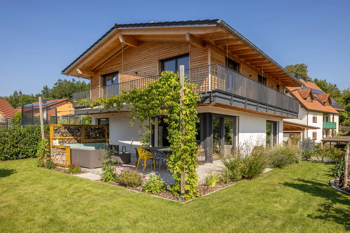 Klassische Einfamilien-Holzhaus mit umlaufendem Balkon in Hanglage. Oben Holzfassade unten verputzt. Gemütliche Terrasse mit Raumteiler aus gestapeltem Stückholz und Kletterwein, der sich zum Balkon hochrankt. © Brunthaler Naturhausmanufaktur
