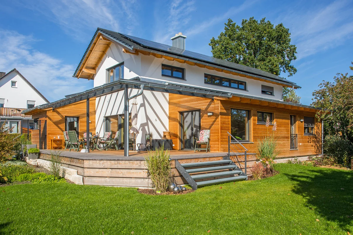 Das Holzhaus ist teilweise verputzt und hat einen überdachten Übereck-Erker mit einer horizontalen Holzfassade. Auf der anderen Seite ist eine überdachte Terrasse mit einer Stahltreppe in den Garten. Das Dach hat graue Schindel. © Brunthaler Naturhausmanufaktur