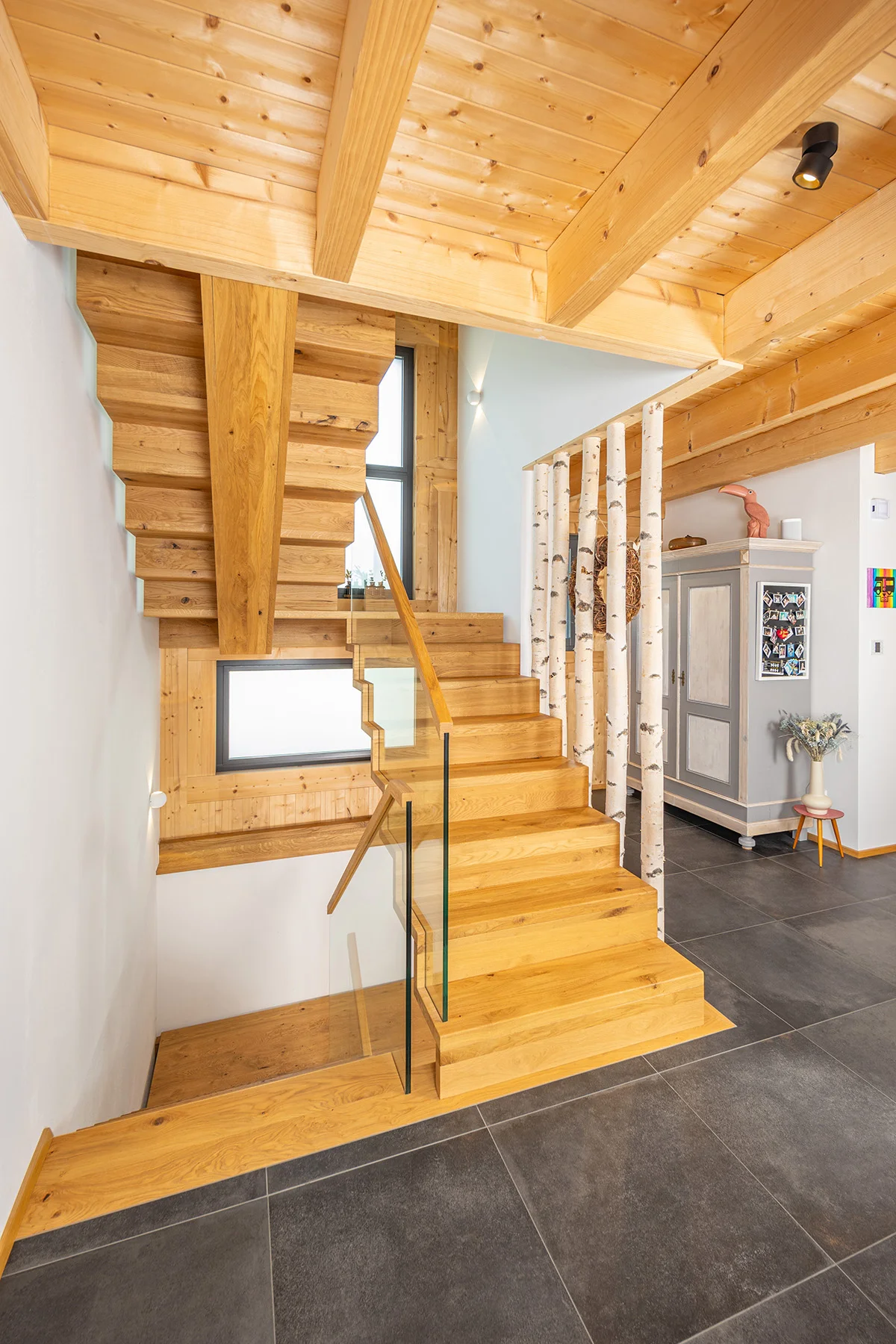 Treppenhaus mit Stufen nach oben und unten. Das Geländer ist modern aus Glas mit Holzgriffen, zusätzlich sind Birkenstämme als Halterung auf anderen Seite eingearbeitet. Man sieht die dunklen großen Bodenfliesen und die Holzdecke des Holzhauses. © Brunthaler Naturhausmanufaktur