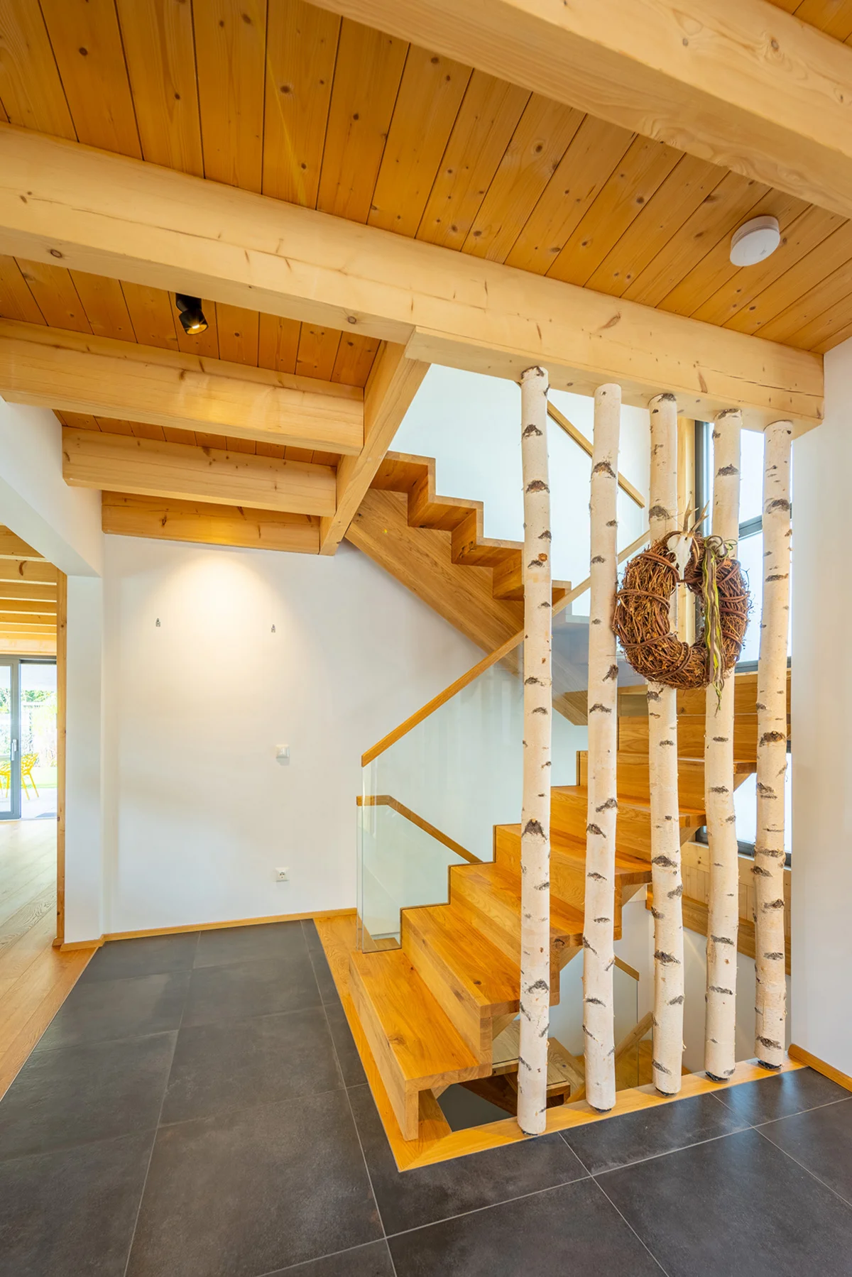 Treppenhaus mit Stufen nach oben und unten. Das Geländer ist modern aus Glas mit Holzgriffen, zusätzlich sind Birkenstämme als Halterung auf anderen Seite eingearbeitet. Man sieht die dunklen großen Bodenfliesen und die Holzdecke des Holzhauses. © Brunthaler Naturhausmanufaktur